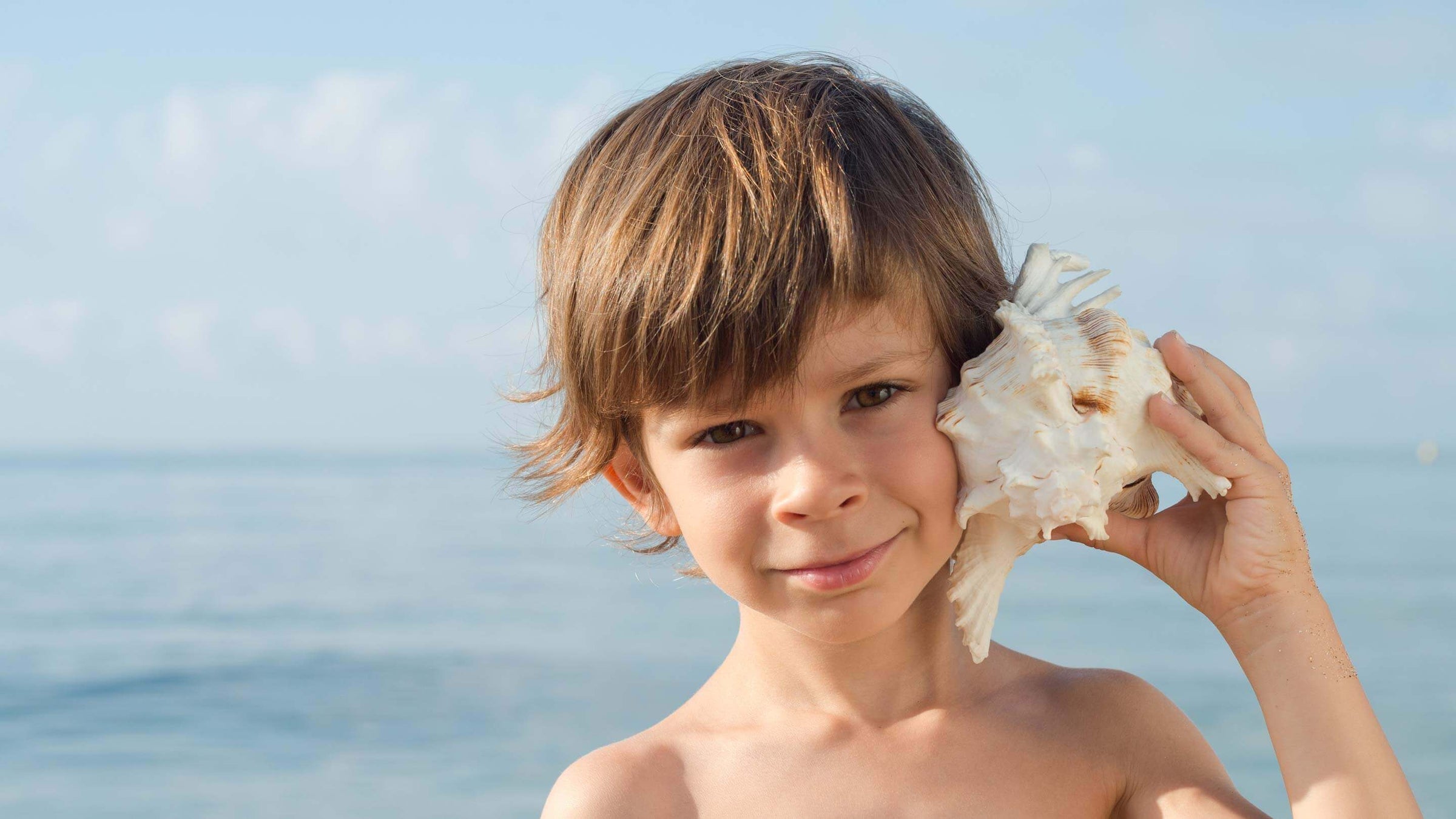 Young boy listening to a conch shell near the ocean.