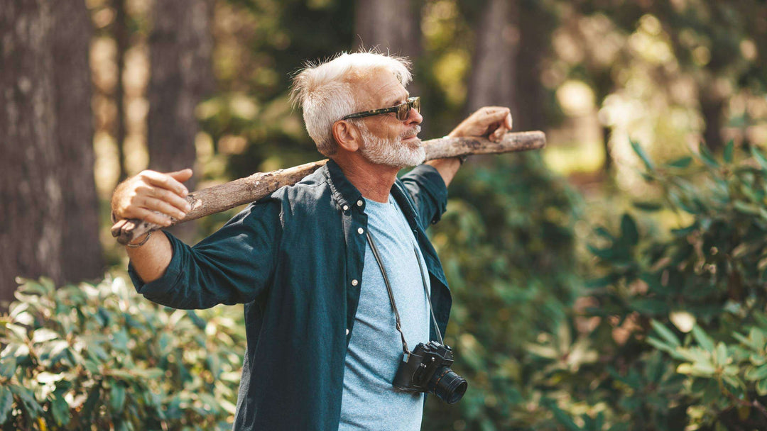 A mature, active, bearded man holds a stick on his shoulders. A pensioner man enjoys life, walking in the woods.