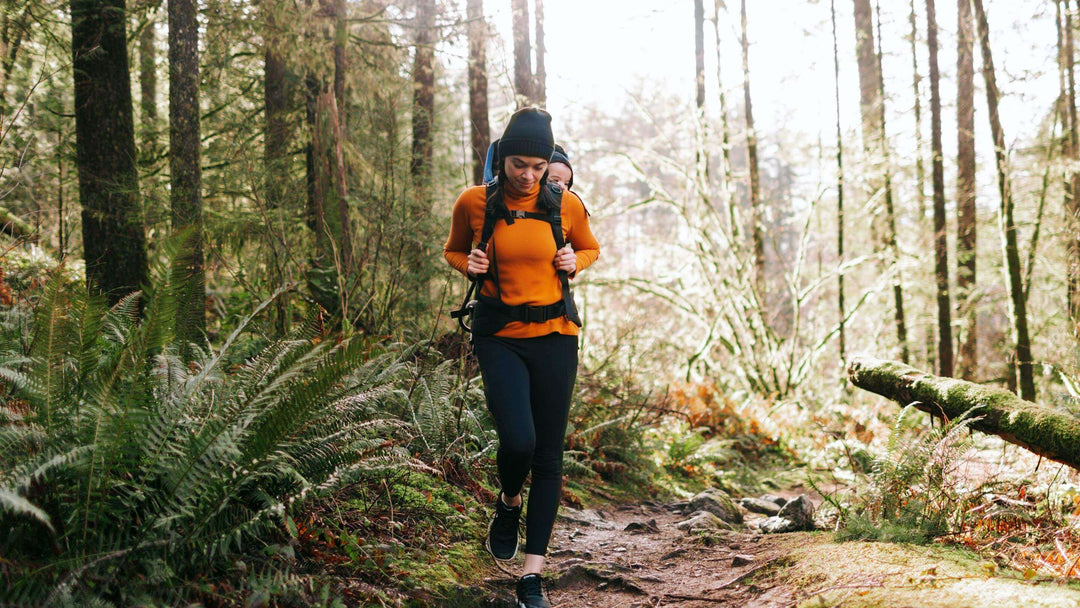 A young woman hiking through the forest with a baby on her back. 