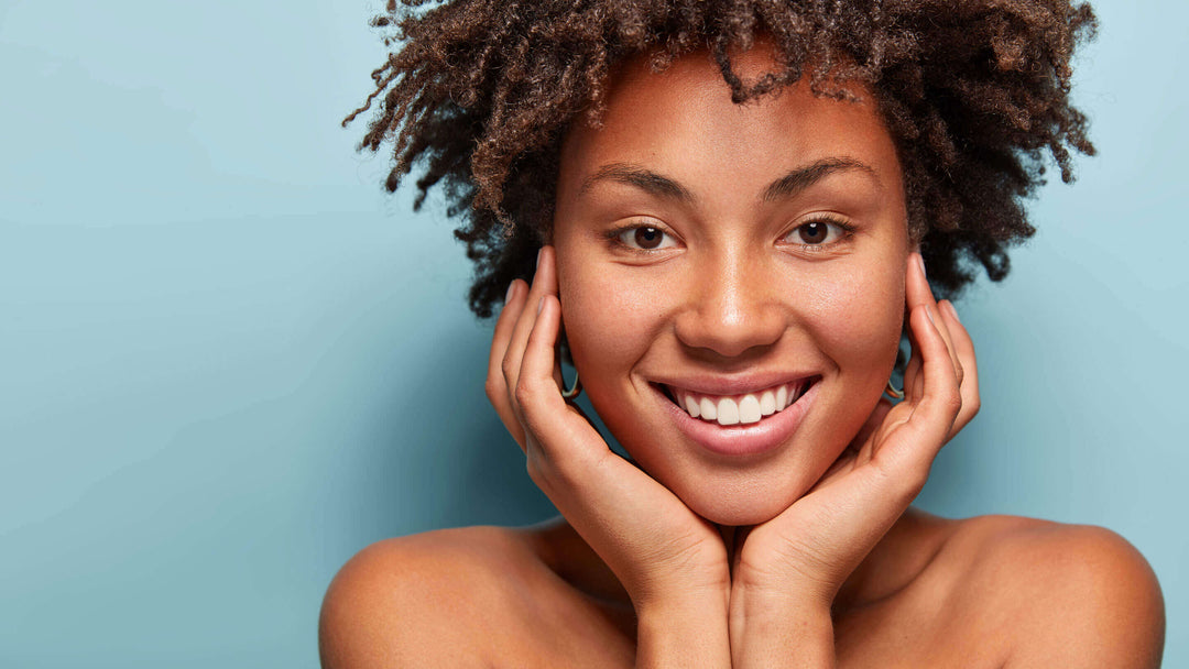 Close up portrait of relaxed black woman has gentle skin after taking shower, satisfied with new lotion, has no makeup, smiles tenderly, shows perfect teeth, stands shirtless against blue background.