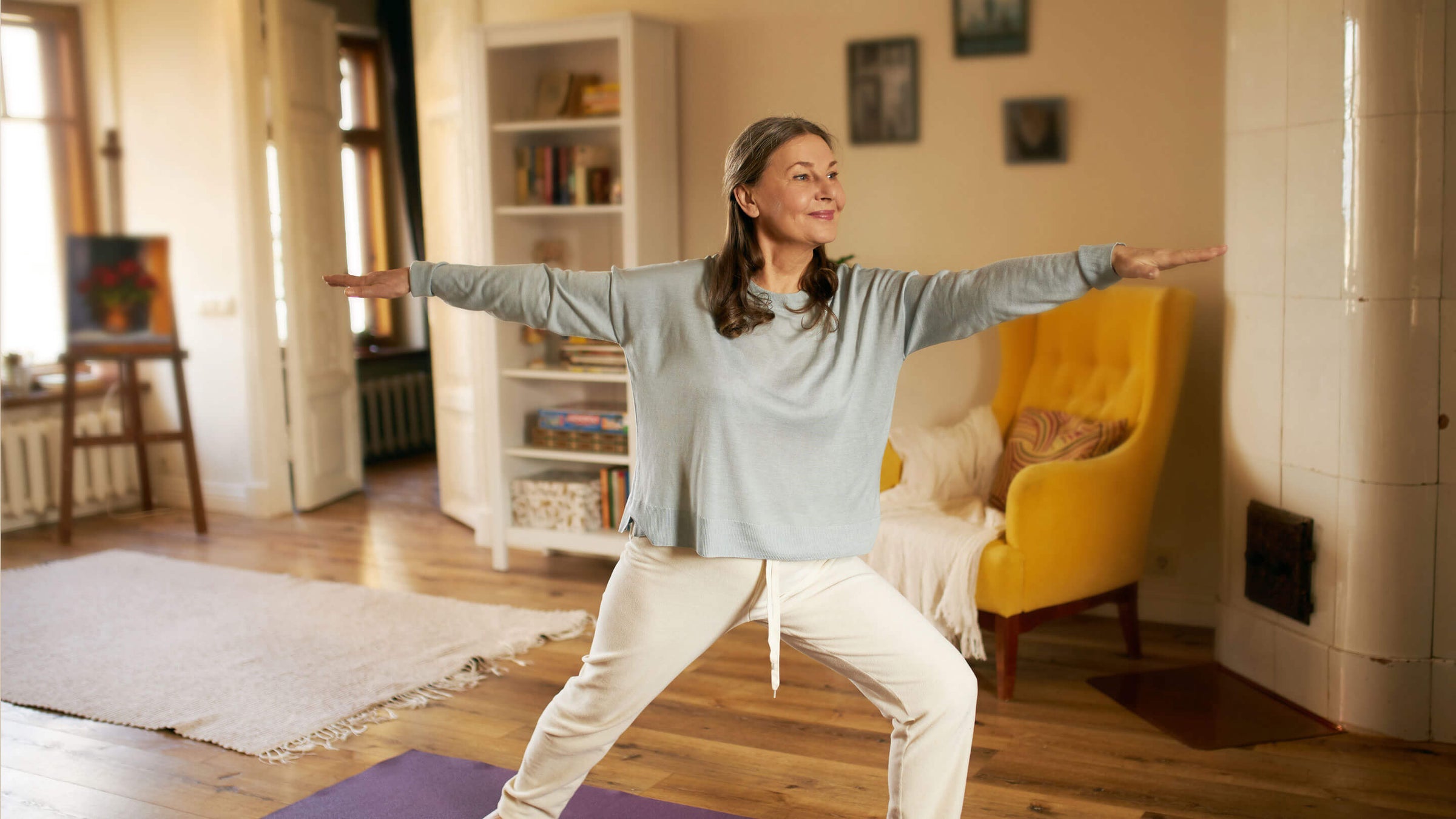 Full length shot of happy energetic mature woman in casual clothes exercising at home because of social distancing, practicing yoga on mat, standing in warrior II pose.